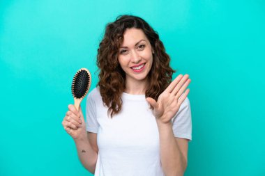 Elinde saç fırçası olan beyaz bir kadın mavi arka planda izole edilmiş, mutlu bir ifadeyle selamlıyor.