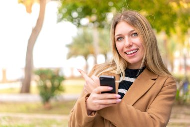 Açık havada cep telefonu kullanan ve arkasını gösteren güzel sarışın bir kadın.