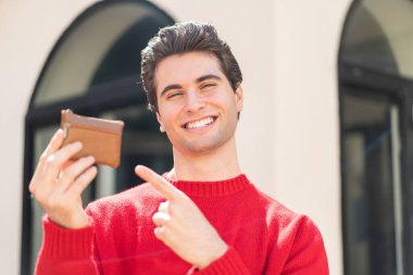 Genç yakışıklı bir adam açık havada bir cüzdan tutuyor ve ona doğrultuyor.