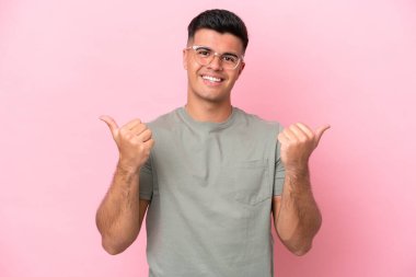 Pembe arka planda izole edilmiş beyaz, yakışıklı bir genç. Baş parmakları havada ve gülümsüyor.