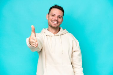 Beyaz tenli yakışıklı adam mavi arka planda izole edilmiş ve iyi bir anlaşma için el sıkışıyor.