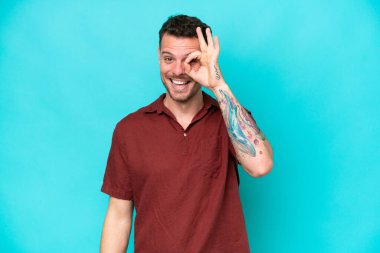 Mavi arka planda izole edilmiş beyaz, yakışıklı genç bir adam parmaklarıyla tamam işareti gösteriyor.