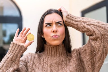 Açık havada Bitcoin tutan genç bir kadın kuşkuları ve kafa karıştırıcı bir ifadeyle