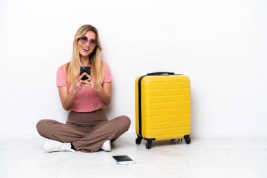 Uruguaylı gezgin kadın, beyaz arka planda izole bir şekilde yerde oturuyor ve cep telefonuyla bir mesaj gönderiyor.