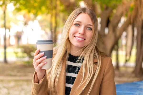 年轻漂亮的金发女人在户外端着咖啡 面带微笑 — 图库照片