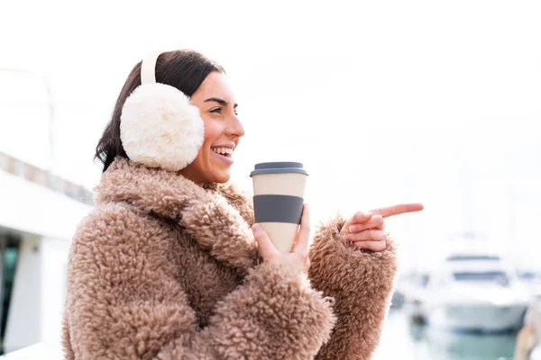 年轻女人穿着冬衣 在户外拿起咖啡 指着旁边介绍自己的产品 — 图库照片