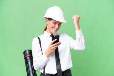 Miğferli, beyaz, genç bir kadın. Arka planda izole edilmiş planlar tutuyor. Telefonu zafer pozisyonunda.