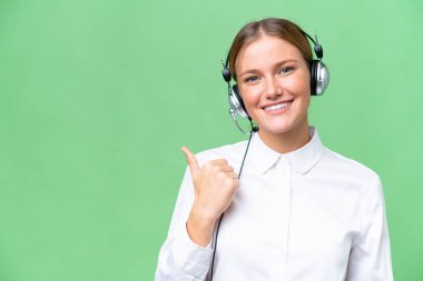 Tele pazarlamacı beyaz kadın izole edilmiş arka planda çalışan bir kulaklıkla bir ürünü sunmak için kenarı işaret ediyor.