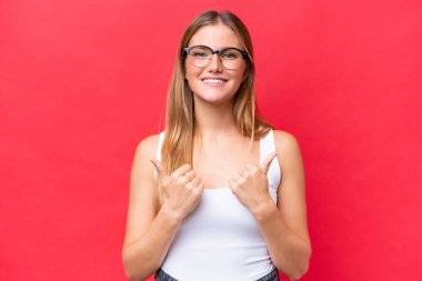 Kırmızı arka planda izole edilmiş genç ve güzel bir kadın. Baş parmakları havada ve gülümsüyor.