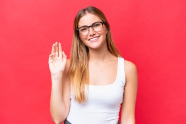 Kırmızı arka planda izole edilmiş genç ve güzel bir kadın. Mutlu bir ifadeyle selamlıyor.