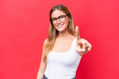 Kırmızı arka planda izole edilmiş genç ve güzel bir kadın parmağını kaldırıyor ve gösteriyor.