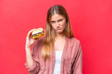 Elinde hamburgerini tutan beyaz bir kadın üzgün bir ifadeyle kırmızı arka planda izole edilmiş.