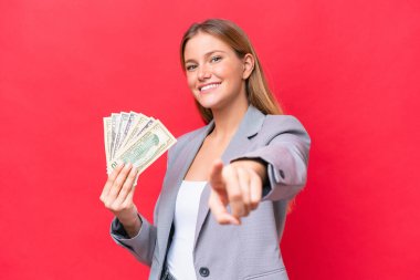 Kafkasyalı genç bir kadın elinde para tutuyor. Kırmızı arka planda izole edilmiş, mutlu bir ifadeyle ön tarafı işaret ediyor.
