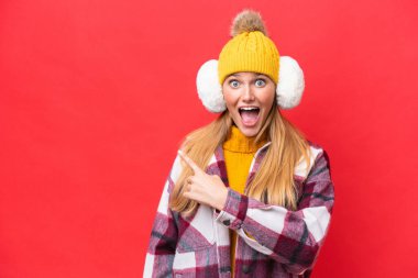 Young beautiful woman wearing winter muffs isolated on red background surprised and pointing side