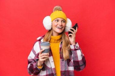 Kırmızı arka planda izole edilmiş kışlık kuklalar takan genç ve güzel bir kadın. Elinde kahve ve cep telefonu var.
