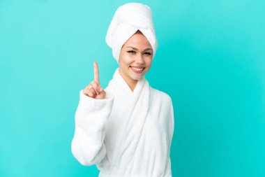 İzole edilmiş mavi arka planda bornozlu genç sarışın kız en iyinin işareti olarak parmağını kaldırıyor.