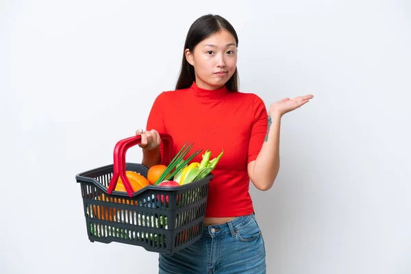 年轻的亚洲女人举着一篮子被白色背景隔离的食物 一边举手一边怀疑 — 图库照片