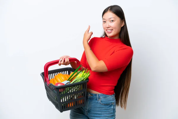 年轻的亚洲女人拿着一个购物篮 里面塞满了被白色背景隔离的食物 然后回头看 — 图库照片