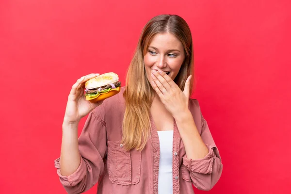 Młoda Kaukaska Kobieta Trzyma Hamburgera Czerwonym Tle Zaskoczeniem Wstrząśniętym Wyrazem — Zdjęcie stockowe