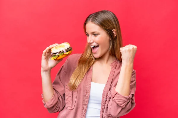 Młoda Kaukaska Kobieta Trzyma Hamburgera Czerwonym Tle Świętując Zwycięstwo — Zdjęcie stockowe