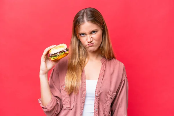 Młoda Kaukaska Kobieta Trzyma Hamburgera Czerwonym Tle Smutnym Wyrazem Twarzy — Zdjęcie stockowe