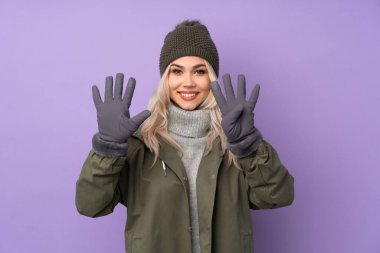 Kış şapkalı genç sarışın kız izole edilmiş mor arka planda parmakları olan dokuzu sayıyor.