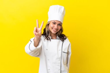 Sarı arka planda izole edilmiş genç Rus şef kız gülümsüyor ve zafer işareti gösteriyor.