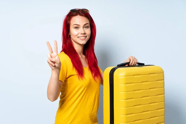 带着一个蓝色背景的行李箱的旅行女孩微笑着 并展示胜利的标志 — 图库照片