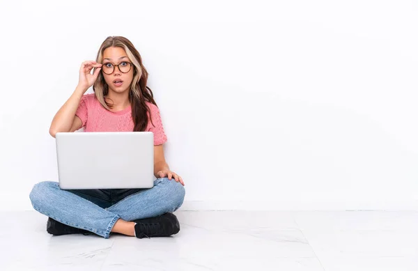 年轻的俄罗斯女孩 带着笔记本电脑坐在地板上 与白色背景隔离 戴着眼镜 惊讶极了 — 图库照片