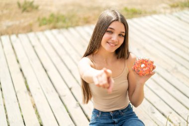 Açık havada donut tutan genç kız kendine güvenen bir ifadeyle seni işaret ediyor.