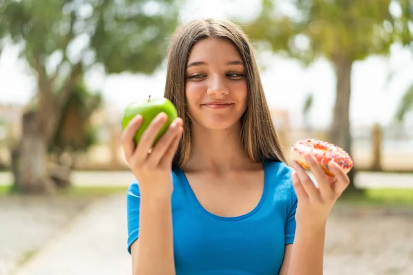 Teenager girl at outdoors holding apple and donut with happy expression