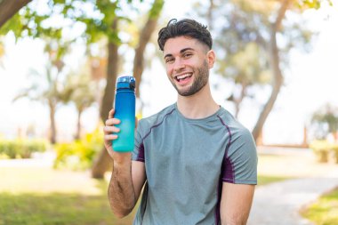 Genç yakışıklı sporcu, elinde bir şişe suyla dışarıda gülümsüyor.