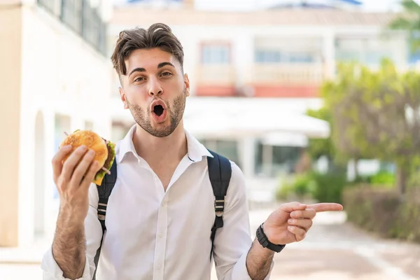 Młody Przystojny Mężczyzna Trzyma Hamburgera Świeżym Powietrzu Zaskoczony Wskazując Bok — Zdjęcie stockowe