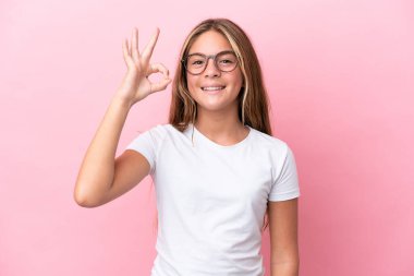Küçük beyaz kız pembe arka planda izole edilmiş gözlüklü ve tamam işareti yapıyor.