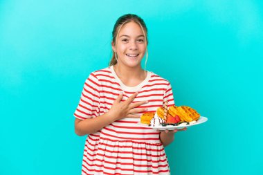 Mavi arka planda izole edilmiş waffleları tutan küçük beyaz kız çok gülümsüyor.