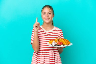 Mavi arka planda izole edilmiş waffle tutan küçük beyaz kız en iyinin işareti olarak parmağını kaldırıyor.