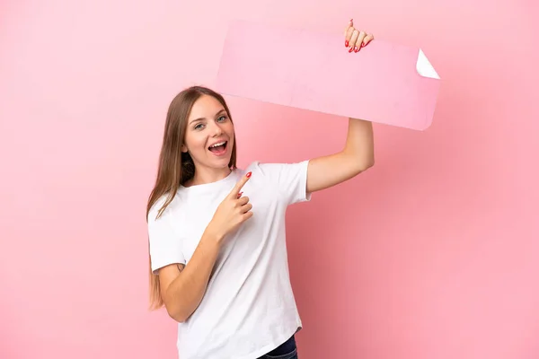 一个年轻的立陶宛女人 被粉色背景隔离 手里拿着一张空标语牌 指着上面 — 图库照片