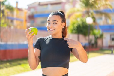 Genç, güzel, esmer bir kadın, açık havada elmalı ve yüzünde sürpriz bir ifade var.