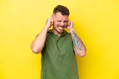 Sarı arka planda izole edilmiş genç beyaz yakışıklı adam hayal kırıklığına uğramış ve kulaklarını kapatmış.