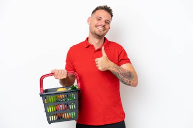 Beyaz arka planda izole edilmiş, elinde yiyecek dolu bir sepet tutan beyaz bir adam baş parmak işareti yapıyor.