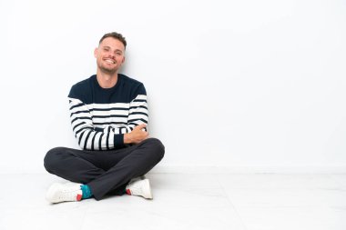 Young man sitting on the floor isolated on white background with arms crossed and looking forward