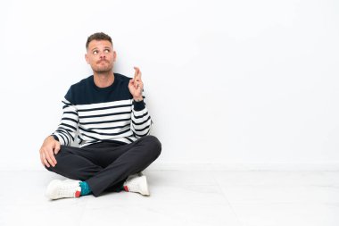 Young man sitting on the floor isolated on white background with fingers crossing and wishing the best