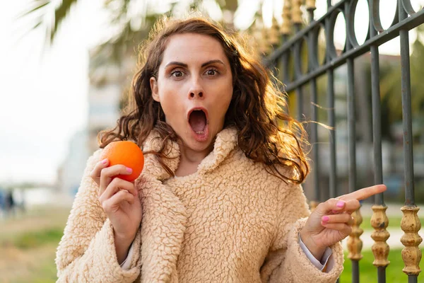 Joven Mujer Caucásica Sosteniendo Una Naranja Aire Libre Sorprendida Señalando —  Fotos de Stock