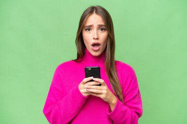 Young beautiful caucasian woman over isolated background looking at the camera while using the mobile with surprised expression