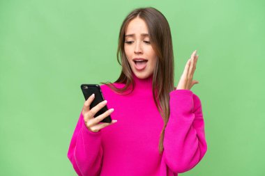 Young beautiful caucasian woman over isolated background looking at the camera while using the mobile with surprised expression