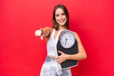 Kızıl arka planda izole edilmiş beyaz bir kadın elinde bir tartı makinesiyle ön tarafı işaret ediyor.
