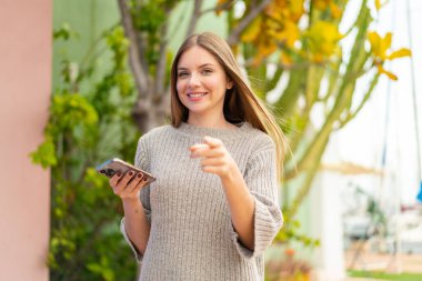 Açık havada cep telefonu kullanan genç, güzel sarışın kadın kendine güvenen bir ifadeyle seni işaret ediyor.