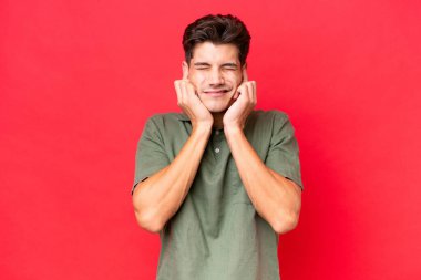 Kızıl arka planda izole edilmiş genç beyaz yakışıklı adam hayal kırıklığına uğramış ve kulaklarını kapatmış.