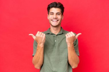 Genç beyaz yakışıklı adam kırmızı arka planda izole edilmiş. Baş parmakları havada ve gülümsüyor.