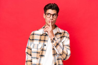 Kırmızı arka planda izole edilmiş beyaz, yakışıklı genç bir adam sessizlik işareti gösteriyor. Parmağını ağzına sokuyor.